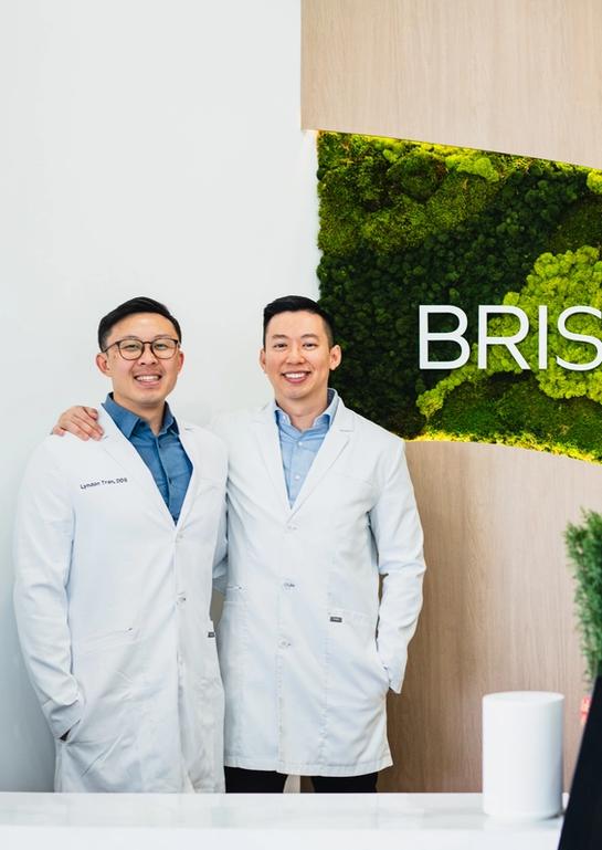 Two smiling men in white lab coats stand in front of a green moss wall sign that reads "BRISTOL MODE".
