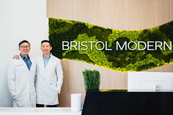 Two smiling men in lab coats stand in a modern reception area with "BRISTOL MODERN" signage on a green moss wall.