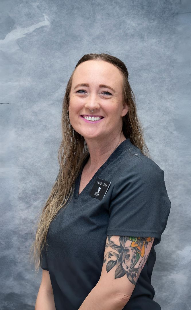 Smiling woman in grey scrubs with an arm tattoo and nose ring.