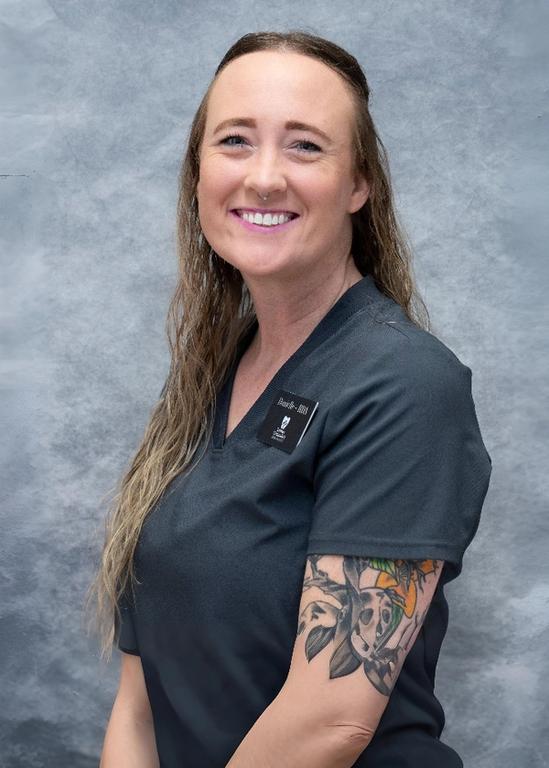 Smiling woman in grey scrubs with an arm tattoo and nose ring.