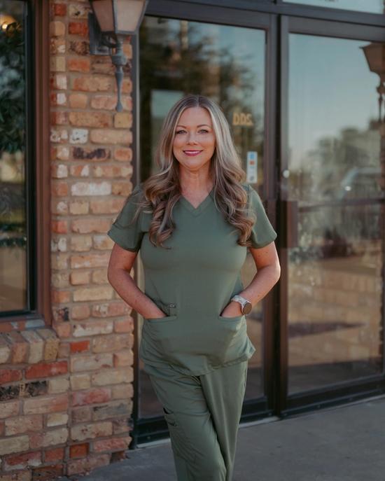 A smiling woman in green scrubs stands outside a dental office.