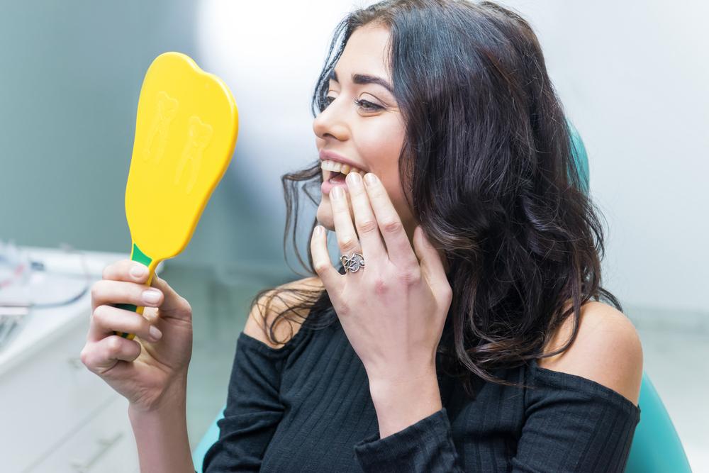 a woman is sitting in a dental chair looking at her teeth in a mirror.