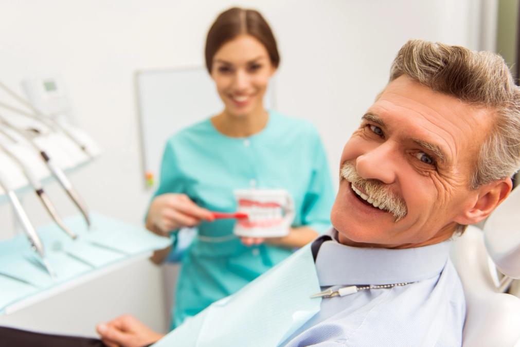 an elderly man is sitting in a dental chair while a dentist shows him how to brush his teeth .