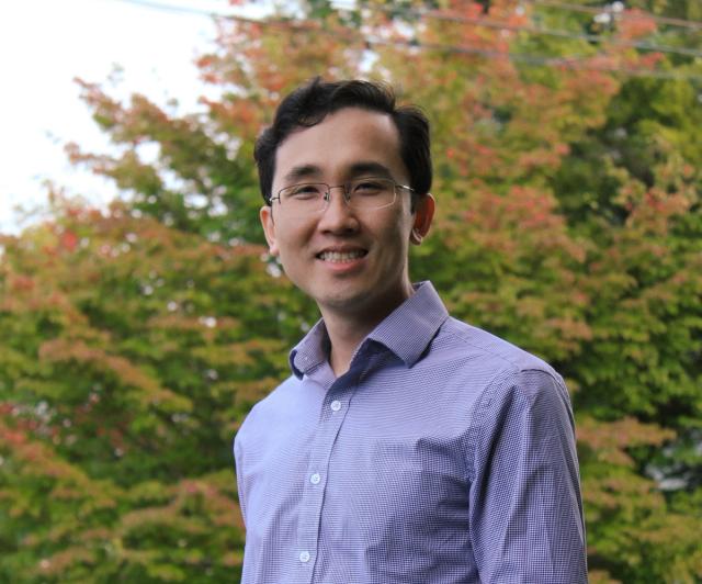 Dr. Anh Duong wearing glasses and a purple shirt is standing in front of a tree .