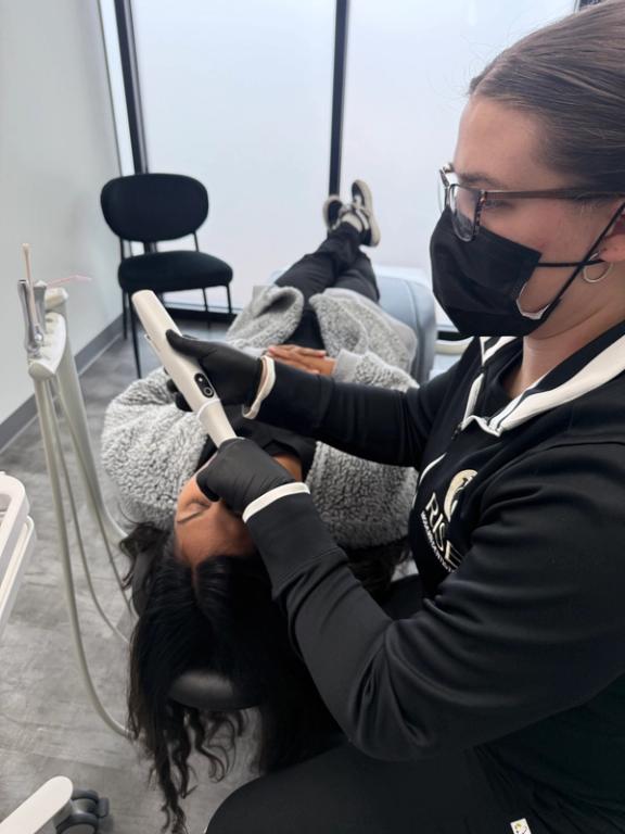 A masked dental professional uses a handheld scanner on a patient's forehead while they recline in a dental chair.