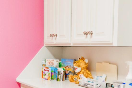A white counter with children's books, a tiger toy, and medical supplies, below white cabinets and next to a pink wall.