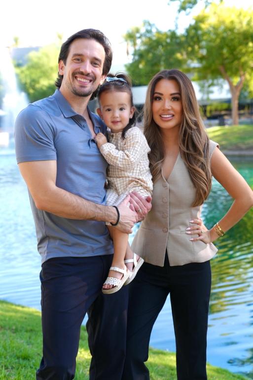 A smiling family of three (man, woman, and young child) poses outdoors by a fountain and water.