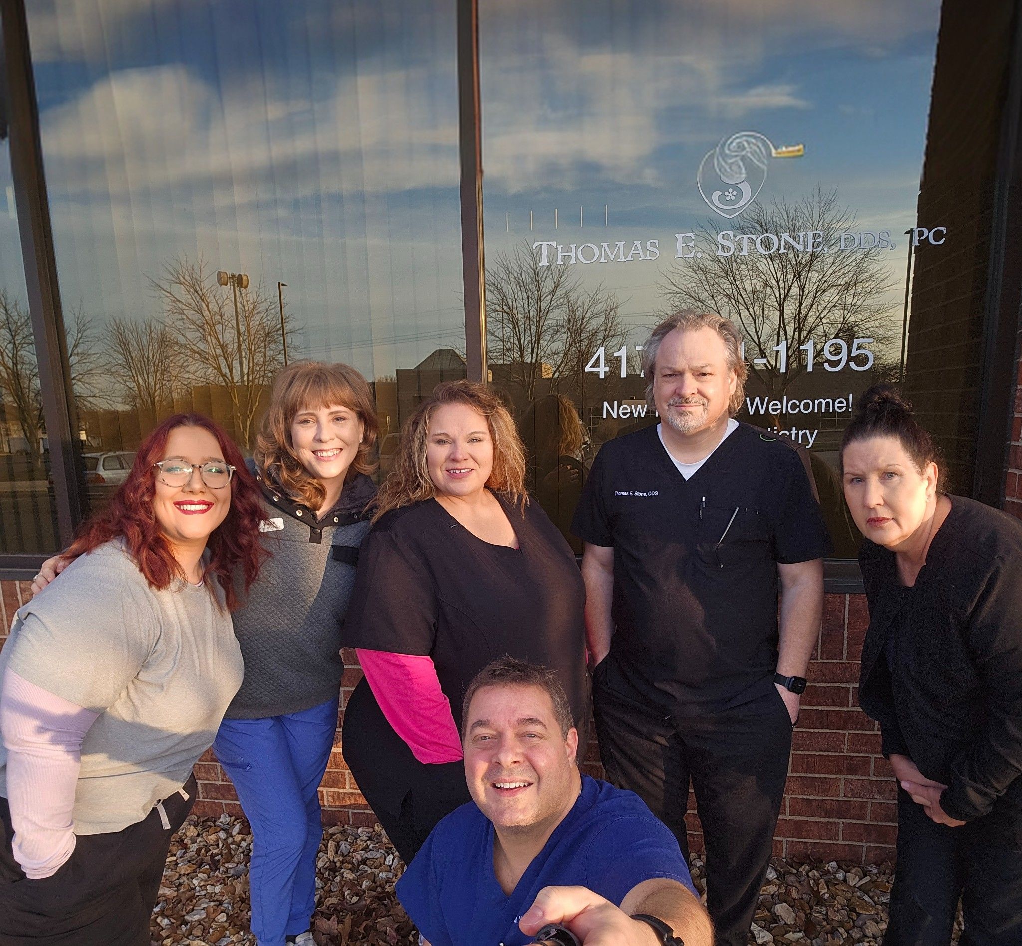 A dental team of five women and one man poses outside Thomas E. Stone, DDS, PC office, one taking a selfie.