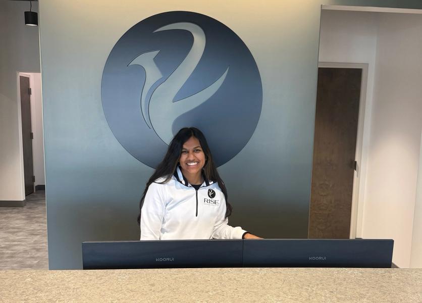 Smiling woman in a RISE shirt behind a counter, with a large RISE logo on the wall.