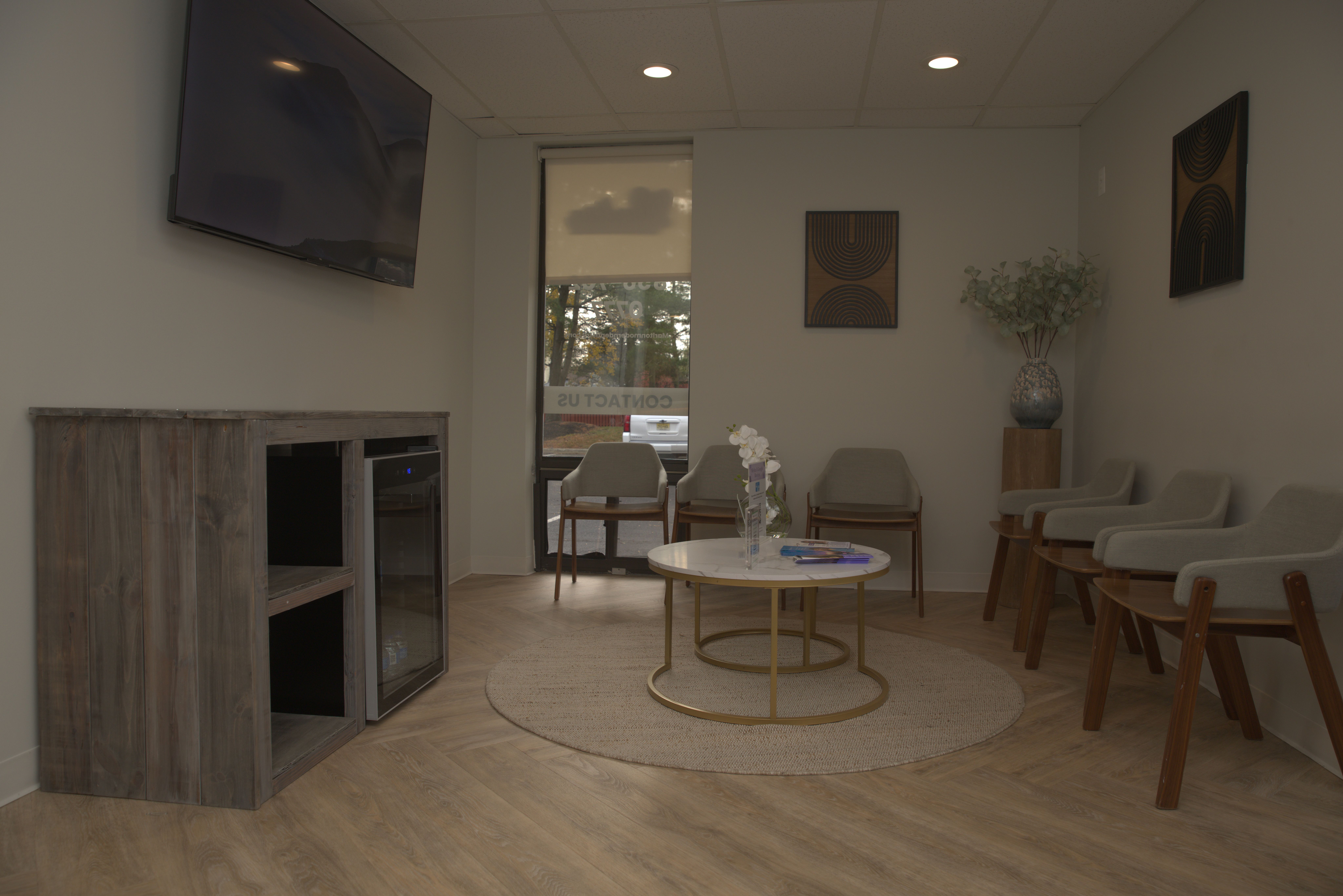 A modern waiting room with light gray chairs, a coffee table, a wall-mounted TV, and a cabinet with a beverage cooler.