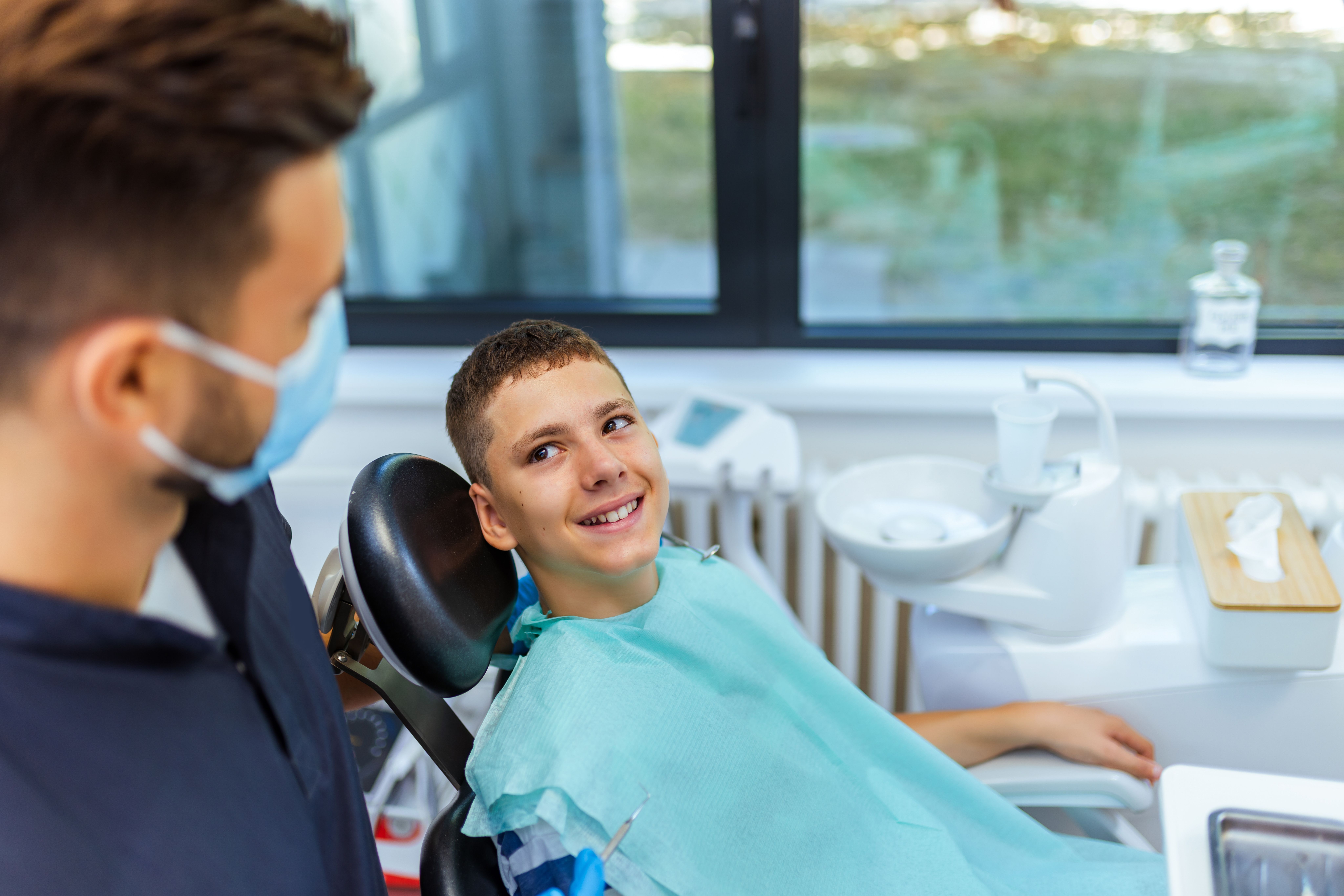 Smiling boy in a dental chair looking at his masked dentist.