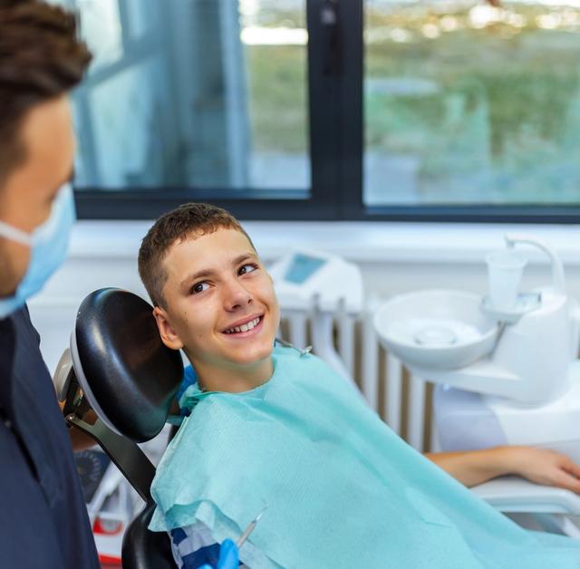 Smiling boy in a dental chair looking at his masked dentist.