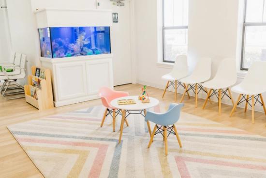 A modern waiting room with a fish tank, a children's play area on a striped rug, and white adult chairs.