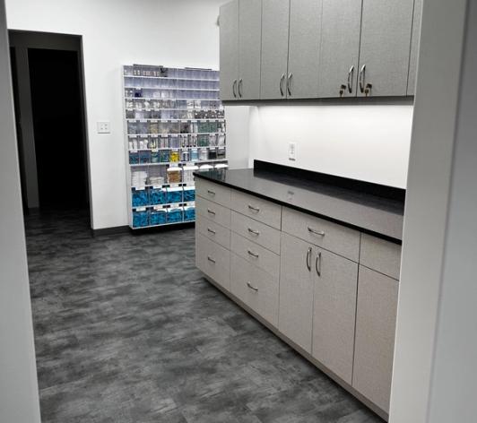 A modern workspace with light gray cabinets, black counters, and a tall shelving unit stocked with supplies.