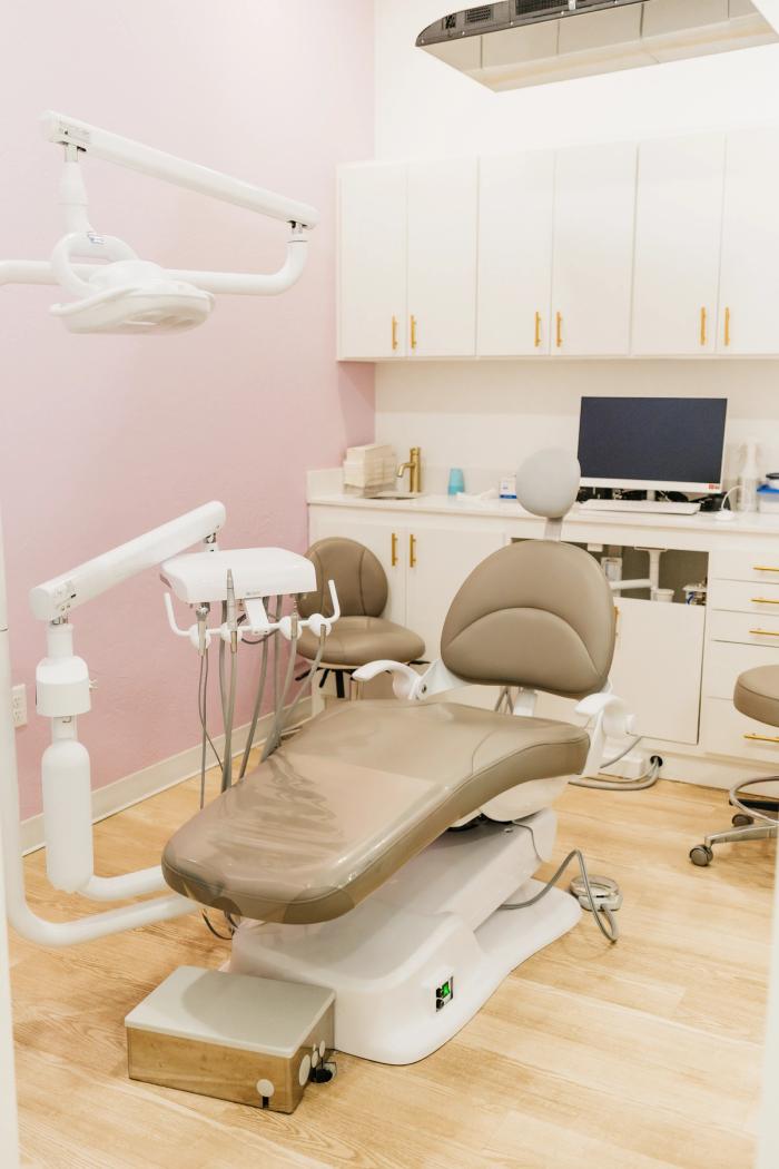 A clean dental examination room featuring a tan chair, white cabinets with gold pulls, a sink, and a pink accent wall.