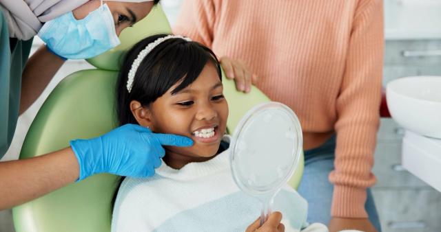 a little girl is sitting in a dental chair looking at her teeth in a mirror .