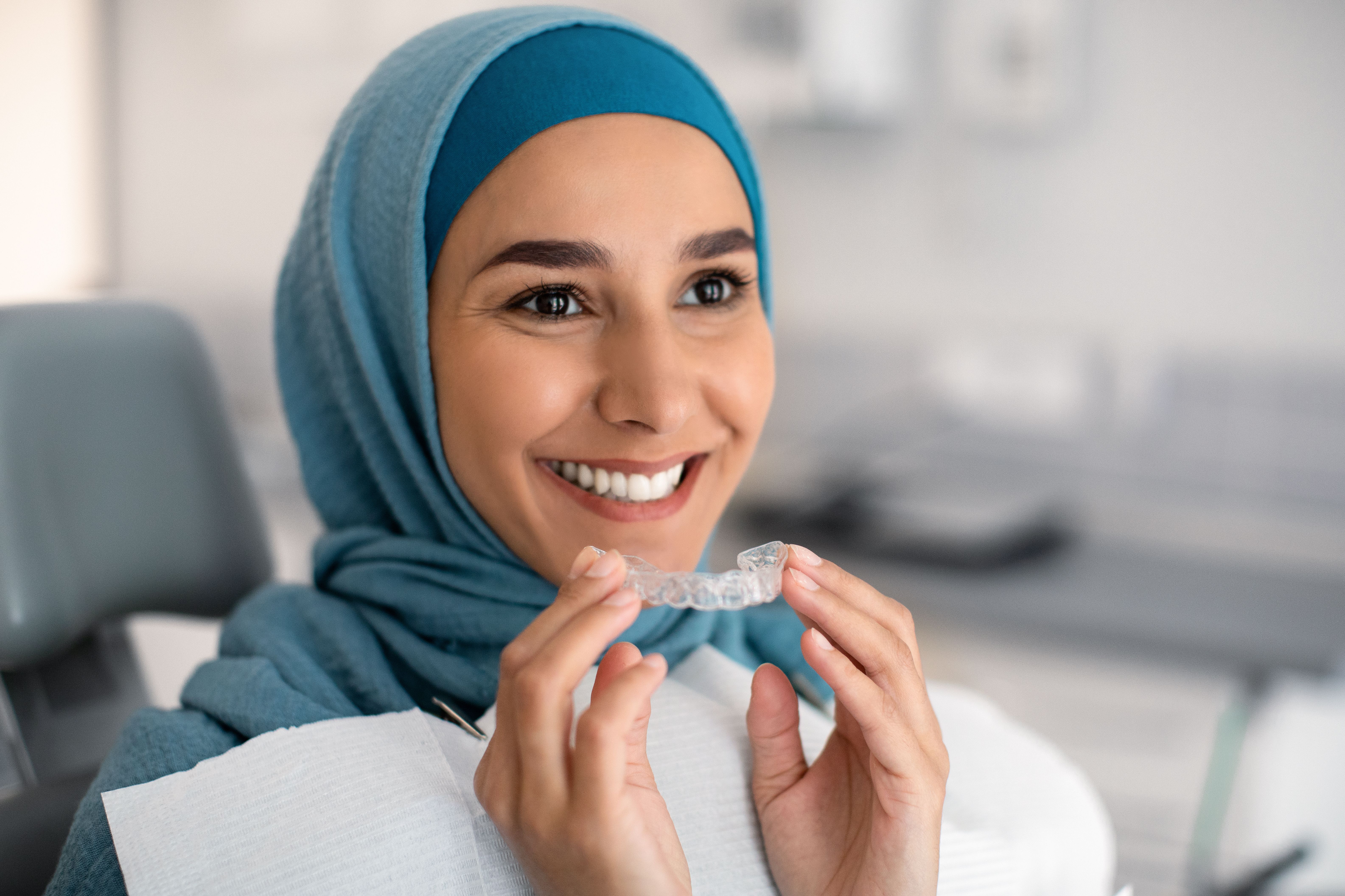 Smiling woman in a hijab holding a clear dental aligner.