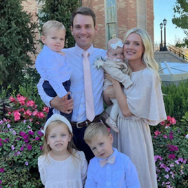 A formal outdoor family portrait of two adults and three young children, including a baby with a feeding tube, surrounded by flowers.
