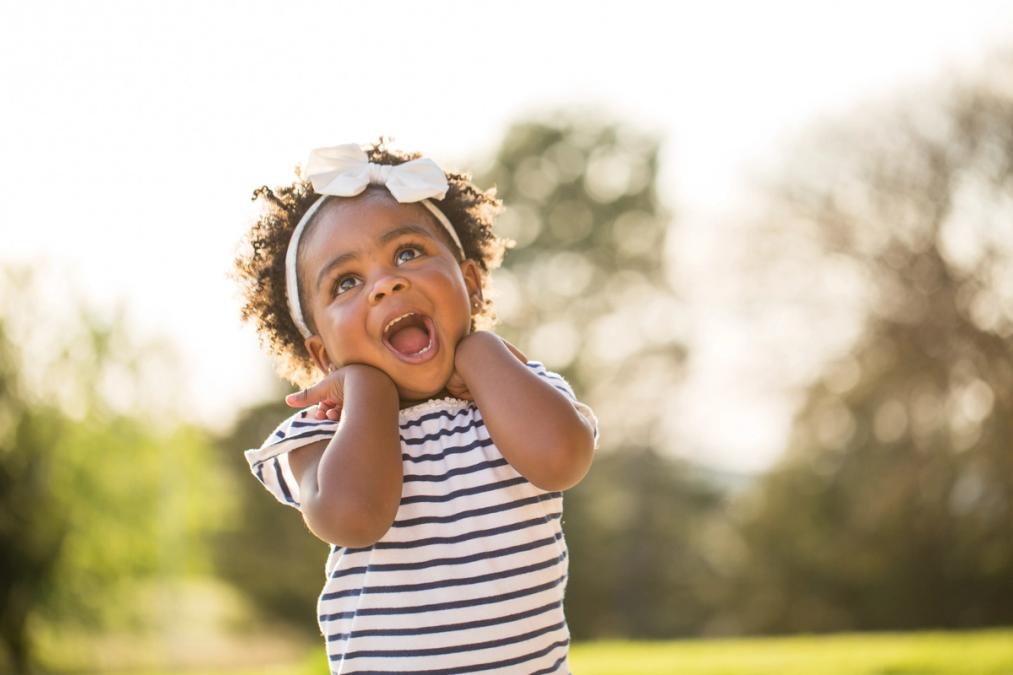 A young child with a white bow in curly hair beams while looking up, hands on cheeks, outdoors.