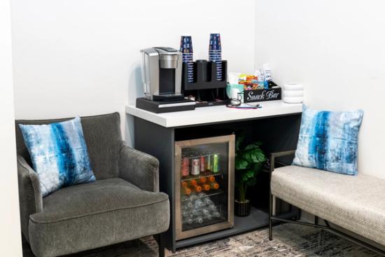Snack and beverage bar in a waiting area with a coffee machine, mini-fridge, and snacks, next to an armchair and bench with blue pillows.