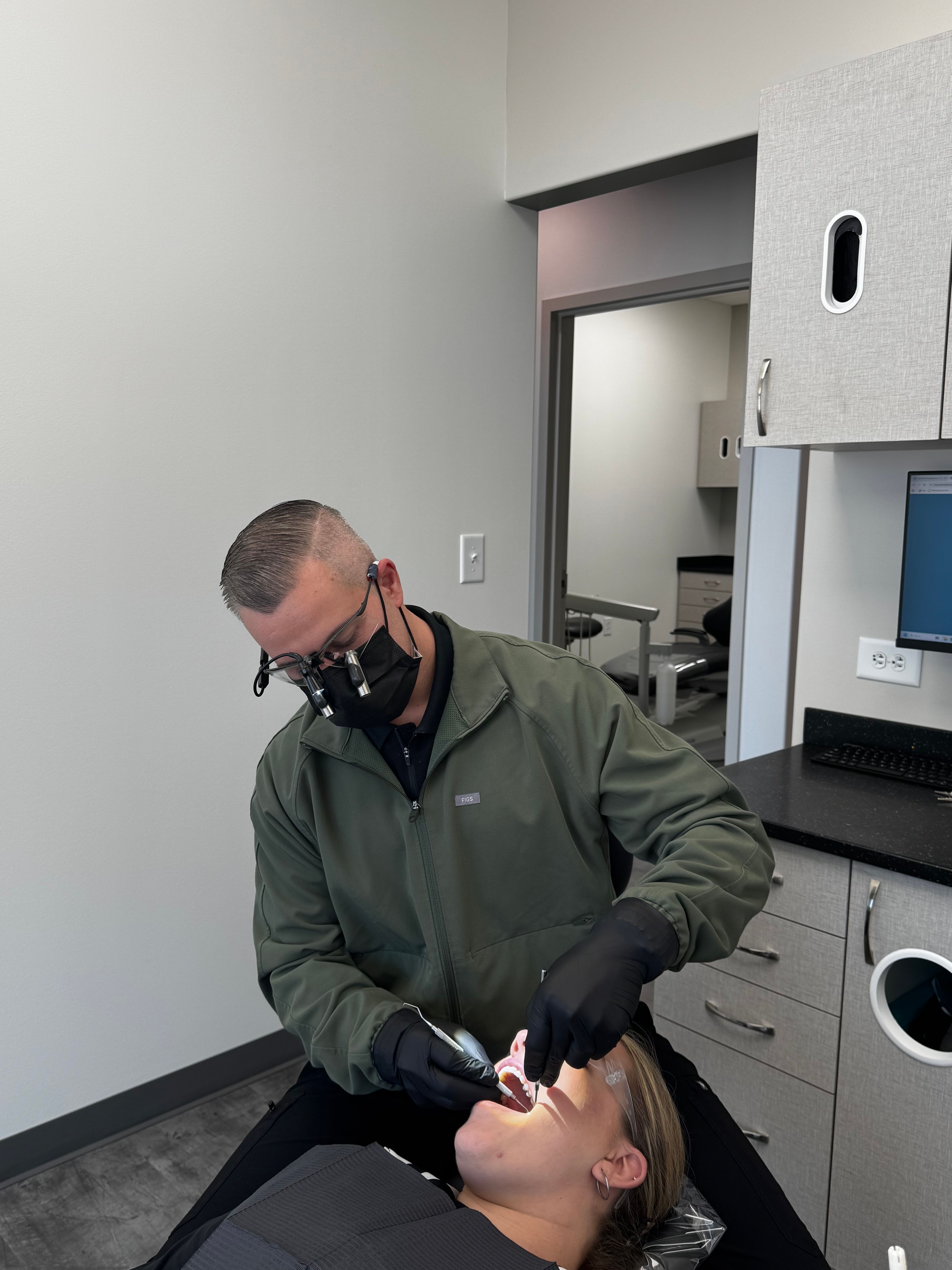 A dentist wearing a mask and loupes examines a patient's mouth.