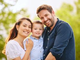 A smiling family of three, a mother, father, and young child, posing outdoors.