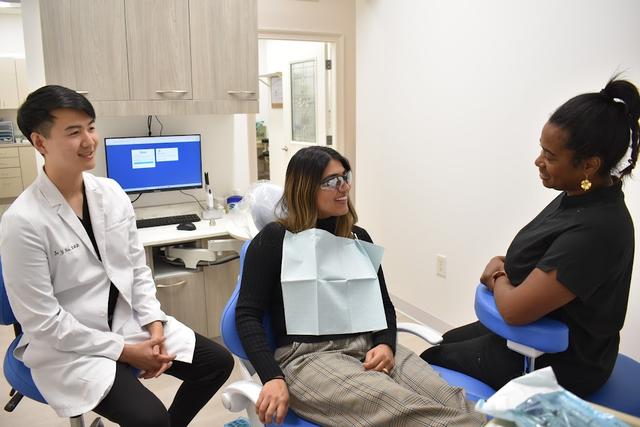 Four smiling individuals, including a man in a white lab coat and a woman in black scrubs, stand in an office reception area.