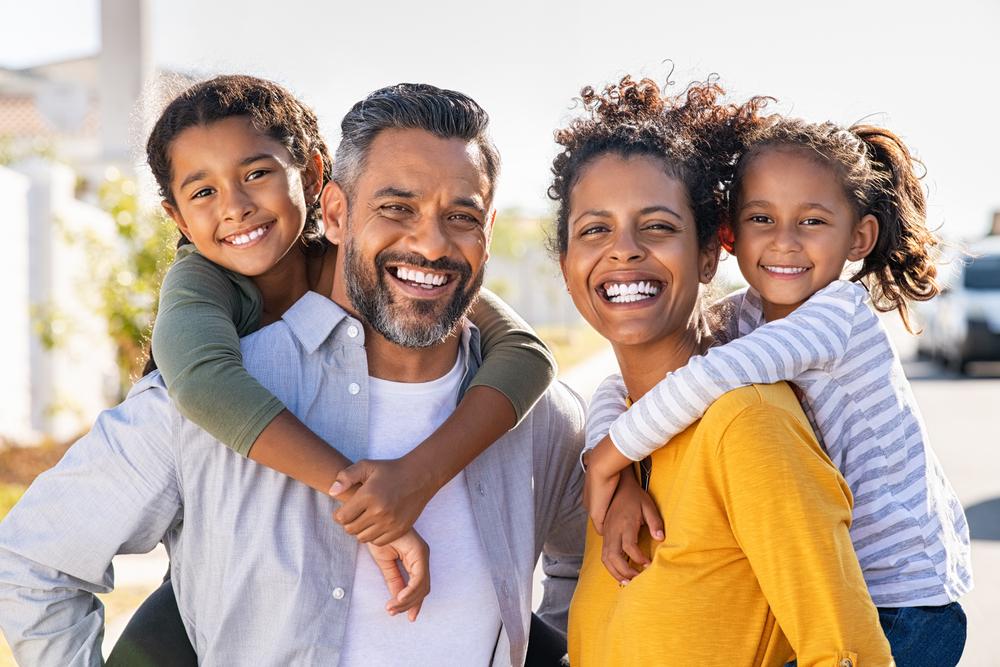 A happy family of four, with parents giving their two daughters piggyback rides, smiling at the camera outdoors.