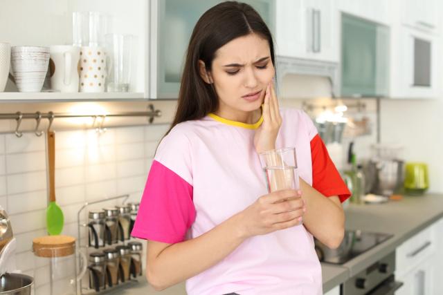 a woman is holding a glass of water and having a toothache in the kitchen .