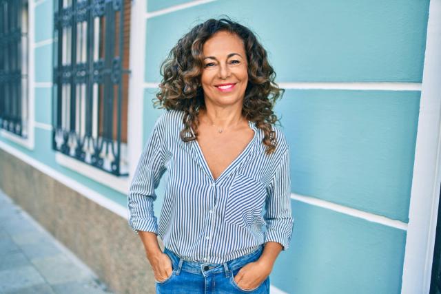 a woman with curly hair is standing in front of a building with her hands in her pockets and smiling .