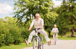 Joyful elderly man riding a bike with legs raised, an elderly woman behind him on a park path.