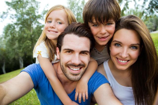 A smiling family of four takes a selfie outdoors in a park.