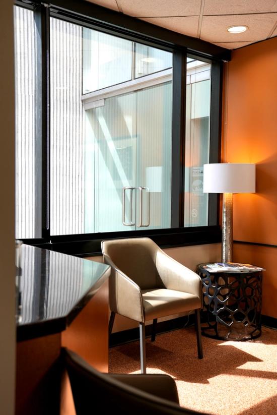 A sunny modern corner with an orange wall, beige armchair, decorative side table with magazines, and a floor lamp beside a large window.