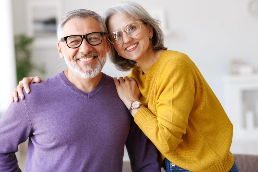 A happy older couple with glasses smiling at the camera, the woman's arm around the man's shoulder.