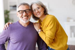 A happy older couple with glasses smiling at the camera, the woman's arm around the man's shoulder.