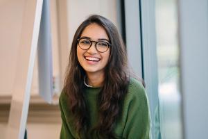 A young woman with long dark hair and glasses smiles broadly.