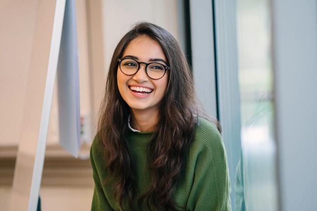 una mujer joven con gafas y un suéter verde sonríe para la cámara.