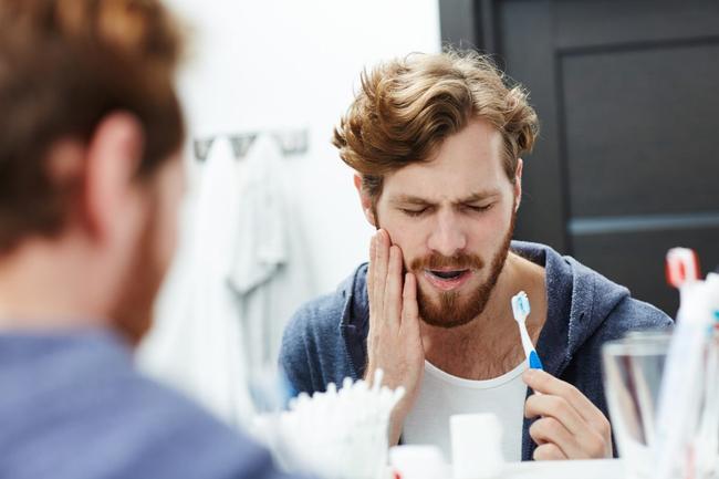 Bearded man holding jaw in pain, with a toothbrush, reflected in a mirror.