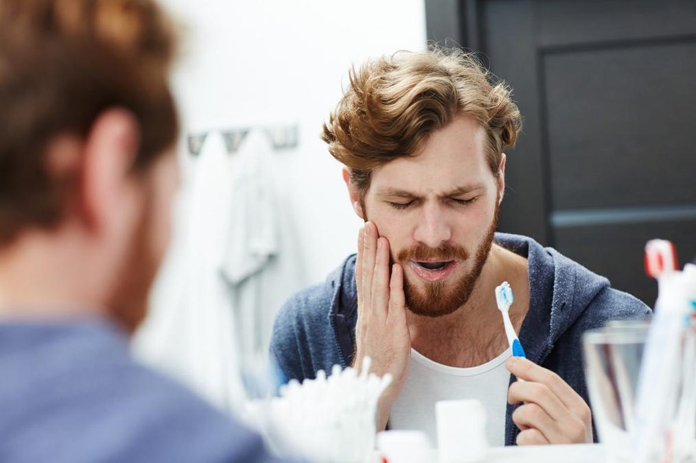 a man is brushing his teeth in front of a mirror while holding his mouth in pain .