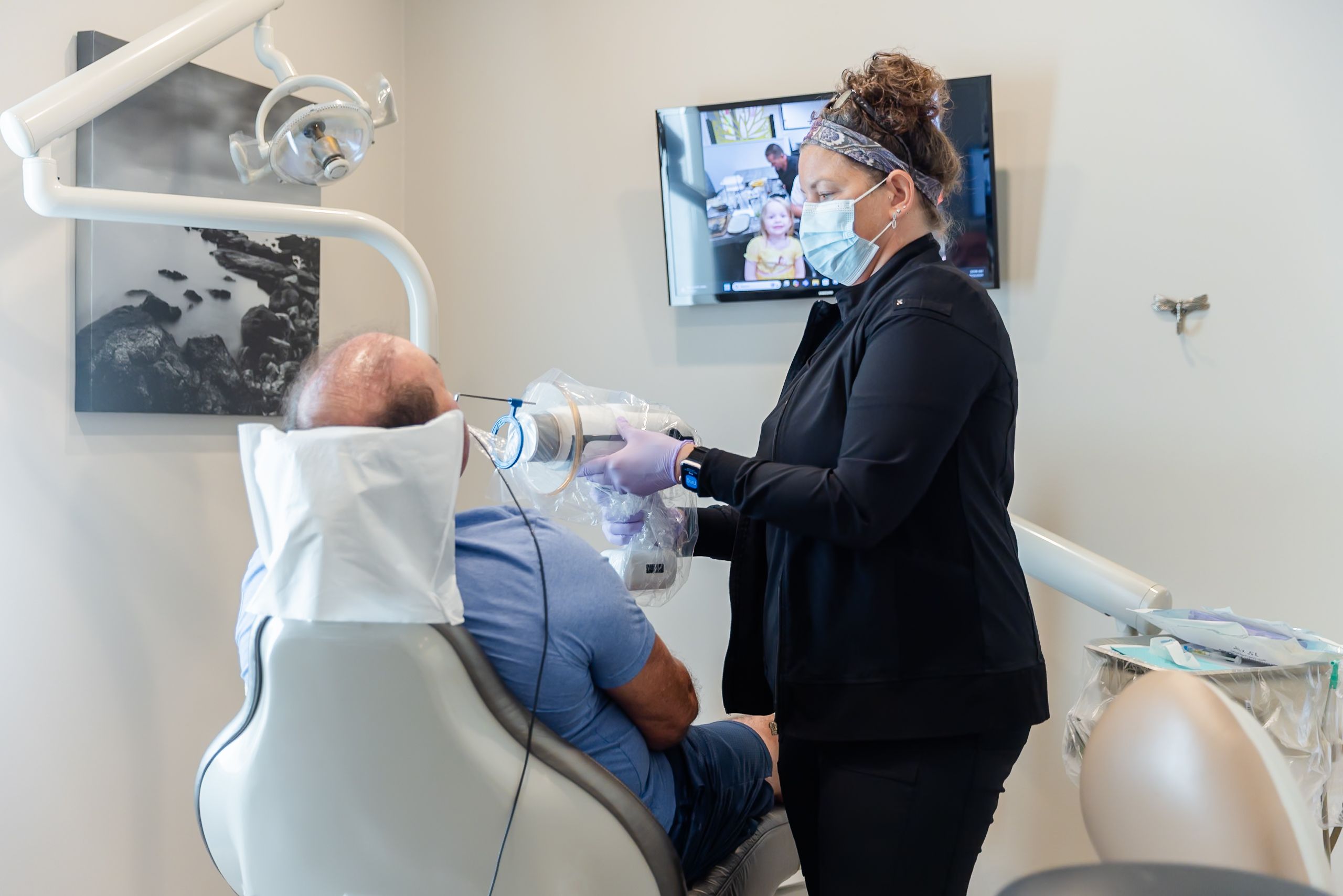 A masked dental professional in gloves uses a portable X-ray machine on a patient.