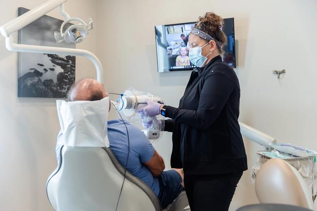A masked dental professional in gloves uses a portable X-ray machine on a patient.