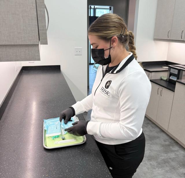 A woman wearing a mask, gloves, and a Rise Rehabilitation Therapy uniform prepares medical supplies on a tray.