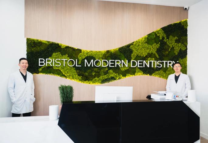 Two dentists in white lab coats stand in the modern reception area of Bristol Modern Dentistry, featuring a green moss wall with the practice name.