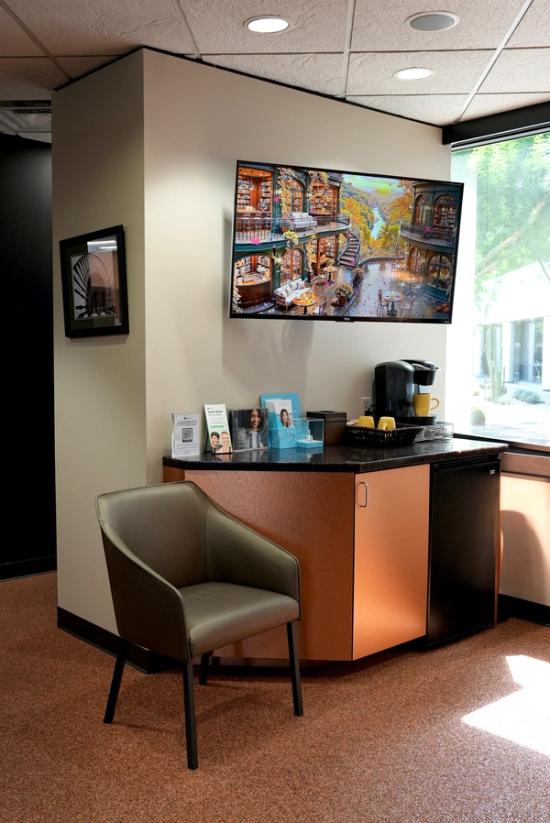 A modern office waiting area featuring a refreshment station with a coffee maker, a TV displaying a vibrant library scene, and a gray leather chair.