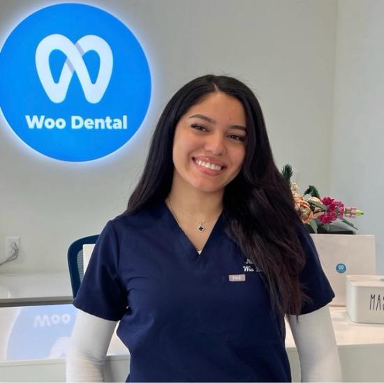 A smiling woman in navy scrubs stands in front of a blue Woo Dental sign.