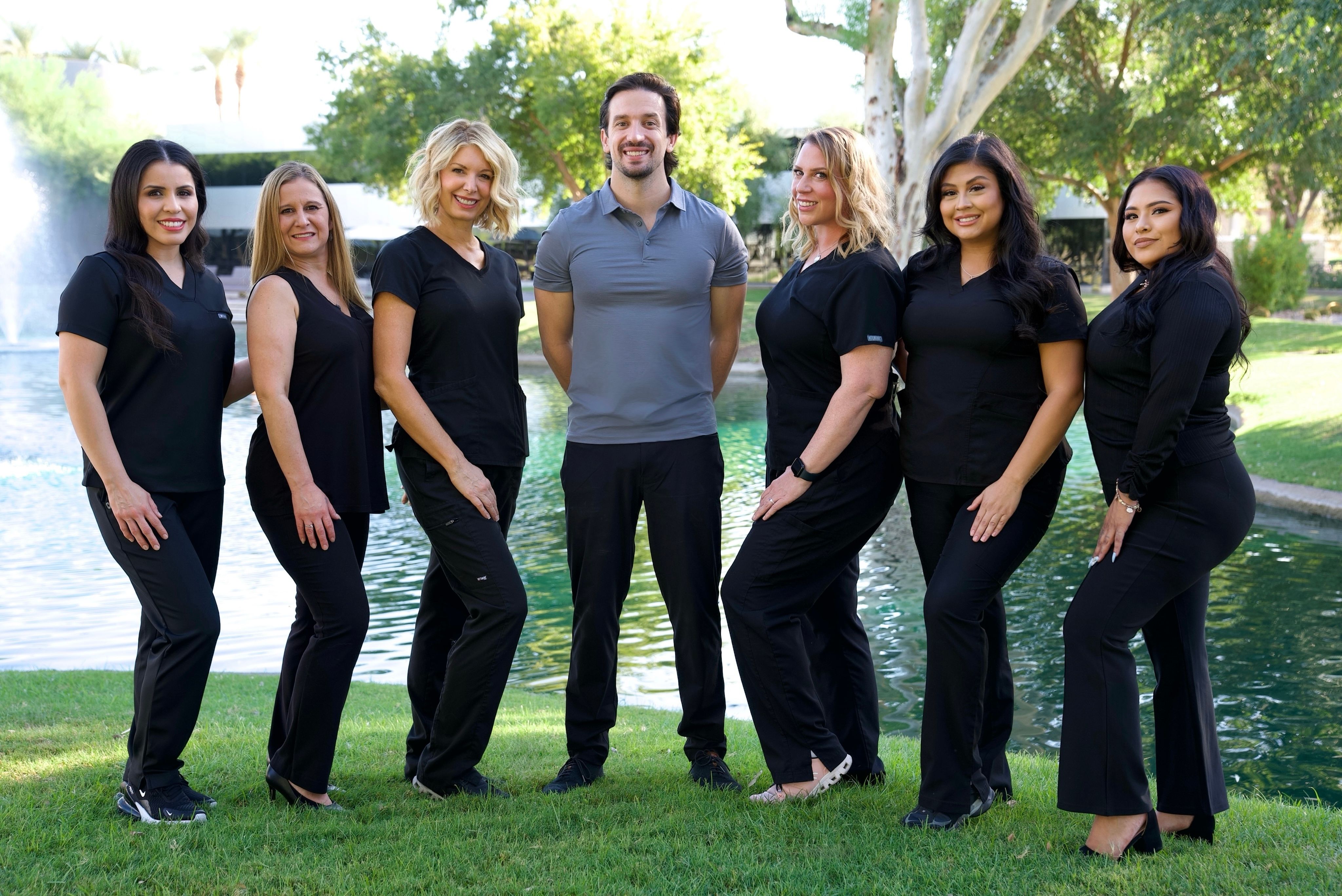A smiling team of eight healthcare professionals (seven women in black scrubs, one man in a grey polo) outdoors by a pond with a fountain.