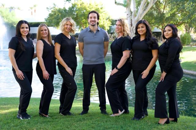 Seven smiling professionals, including one man and six women, stand by an outdoor pond with a fountain.