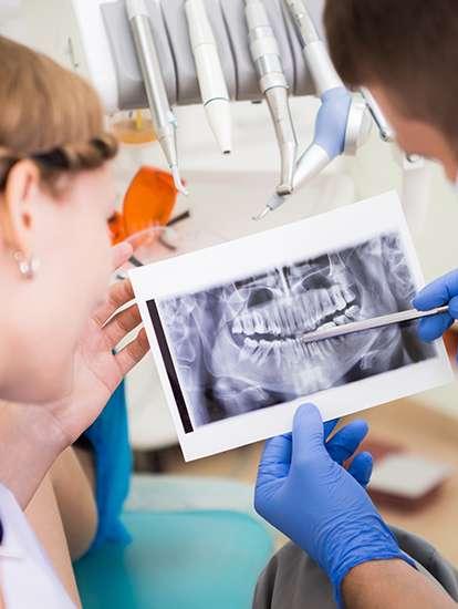A dentist points to a dental X-ray as a patient looks on, with dental tools in the background.
