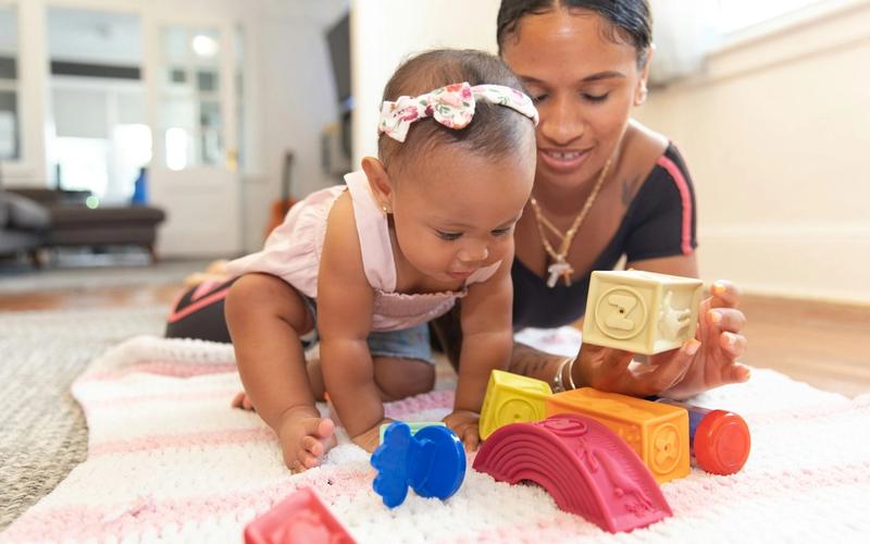 A mother sitting on a blanket playing with toys alongside her baby