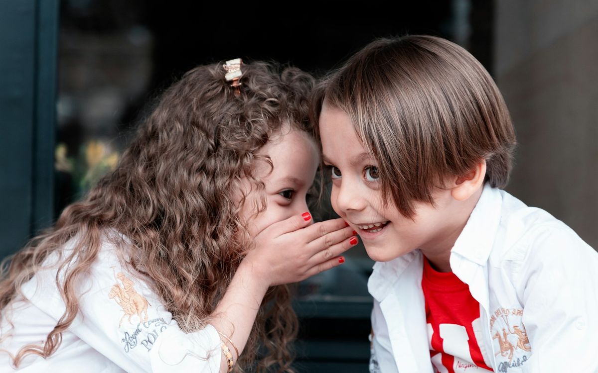 A young girl whispering a secret into a smiling boy’s ear